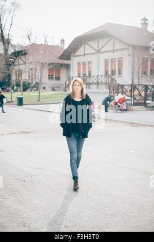 Jeune belle blonde sportive hipster femme marche dans la ville, les mains en poche, donnant sur la droite - porter un jean bleu et manteau noir - jeunes, 20 s, insouciance concept Banque D'Images