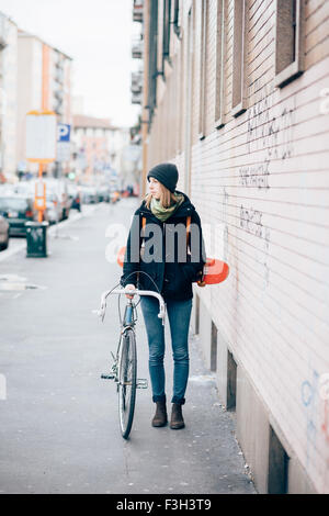 Belle jeune femme blonde sportive hipster marcher dans la ville tenant un vélo et un skate, donnant sur la gauche - concept sportif Banque D'Images