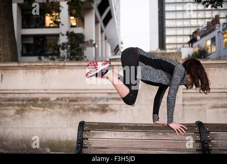 Jeune femme sautant au-dessus de banc de parc en ville Banque D'Images