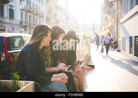 Trois jeunes femmes assises using digital tablet on city street Banque D'Images