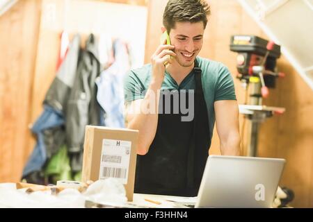 Jeune homme en atelier standing at desk talking on telephone looking at laptop Banque D'Images