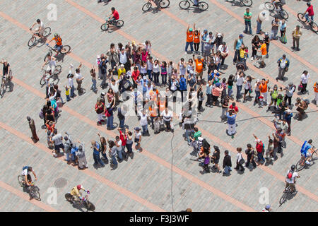 Une prise de vue aérienne au-dessus de la tête capture un grand groupe de personnes rassemblées sur une place pavée avec un motif quadrillé de carreaux gris clair et brun rougeâtre. De nombreux individus dans le groupe central élèvent leur Banque D'Images