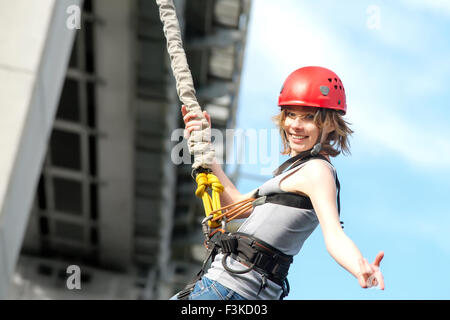 Belle jeune femme dans un casque accroché sur une corde après le saut à l'élastique contre le ciel Banque D'Images