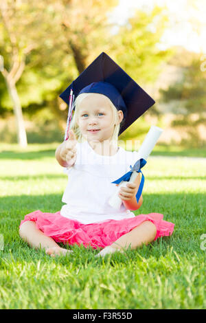 Cute Little Girl In Grass Wearing Graduation Cap Holding Diploma avec ruban. Banque D'Images