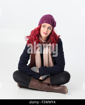 Jolie fille aux cheveux rouges portant manteau d'hiver, bonnet et écharpe en laine violette, de s'asseoir. Isolé sur fond blanc. Banque D'Images