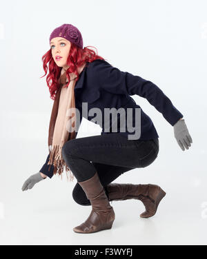 Jolie fille aux cheveux rouges portant manteau d'hiver, bonnet et écharpe en laine violette, accroupi. Isolé sur fond blanc. Banque D'Images