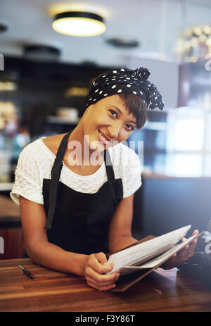 Smiling young coffee house propriétaire debout derrière le comptoir dans un tablier, se penchant en avant pour offrir le menu avec un accueil chaleureux Banque D'Images