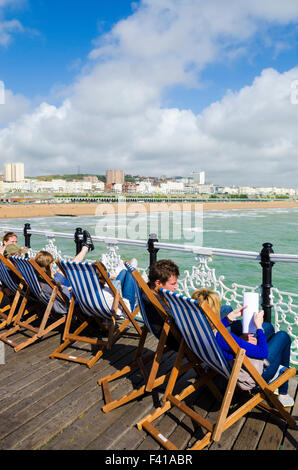 Les gens assis dans des chaises longues sur la jetée de Brighton avec vue sur la mer et la plage de Brighton. East Sussex, Angleterre. Banque D'Images