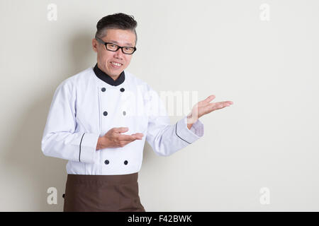 Portrait de 50s mature Asian male chef en uniforme montrant quelque chose, debout sur fond uni avec ombre, copiez l'espace. Banque D'Images