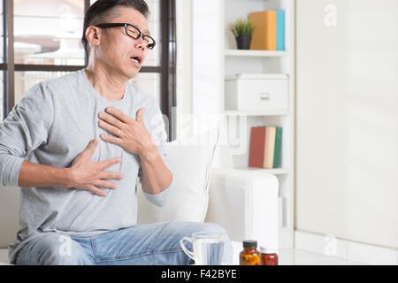 Portrait de 50s occasionnels mature Asian man heartburn, agissant sur poitrine avec expression douloureuse, assis sur le canapé à la maison, des médicaments Banque D'Images