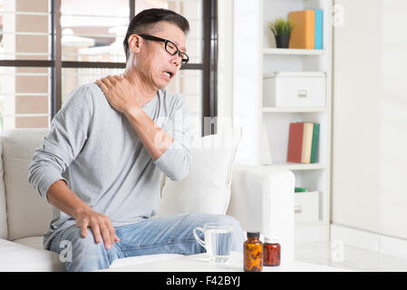 Portrait de 50s occasionnels mature Asian man la douleur de l'épaule, agissant sur le cou avec expression douloureuse, assis sur le canapé à la maison, Medici Banque D'Images