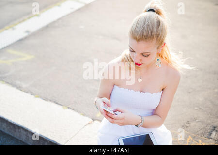 Young handsome young blonde woman long cheveux raides en utilisant des dispositifs technologiques comme smartphone ou tablette - le multitâche, Banque D'Images