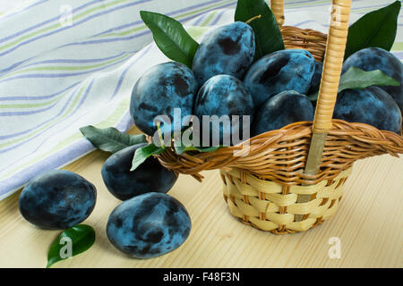 Les prunes mûres dans de petites feuilles vertes panier en osier sur le linge de table en bois avec des serviettes à rayures. Selective focus Banque D'Images