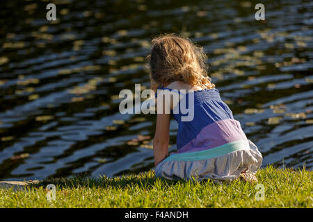 Jeune fille avec des cheveux longs assis avec dos à la caméra sur l'herbe au bord de l'eau Banque D'Images