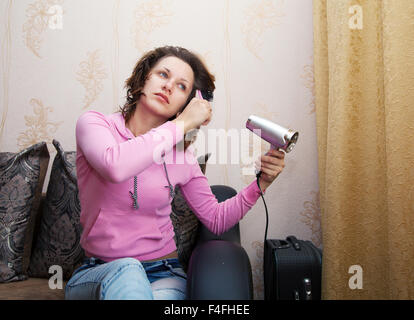 Jeune femme brune en blouse rose et bleu jeans sèche le sèche-cheveux assis sur le canapé Banque D'Images