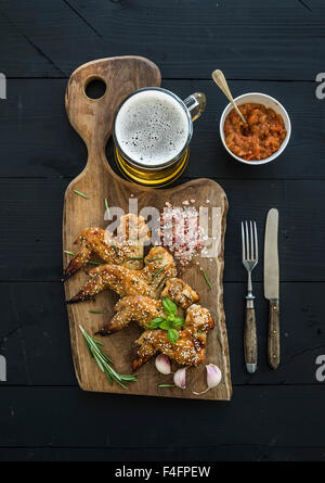Ailes de poulet frit sur du conseil rustique, épicé de la sauce tomate, les herbes et mug de bière légère sur fond de bois noir, top vie Banque D'Images