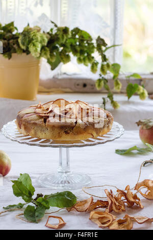 Tarte aux pommes faite maison sur son socle en verre avec des fruits frais et secs, les pommes de table avec nappe blanche. Avec fenêtre rustique au bac Banque D'Images