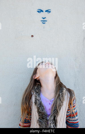 Petite fille à la recherche d'une photo d'un visage peint sur un mur Banque D'Images