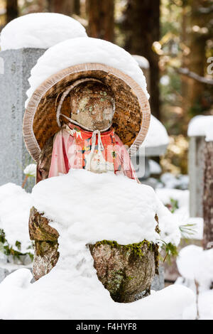 Le Japon, Koyasan, Okunoin cemetery. Assis en pierre couverte de neige-Jizo bosatsu statue bouddhiste, rouge et chapeau de paille conique de soumission. L'hiver Banque D'Images