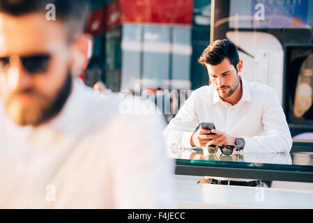 Beau jeune homme moderne cheveux noirs, assis sur un comptoir, à l'aide d'un smartphone à l'écran vers le bas - travail, affaires, technologie concept Banque D'Images