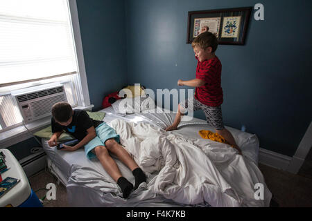 Un jeune garçon se penche sur sa tablette alors que son jeune frère danses complètement sur son lit Banque D'Images