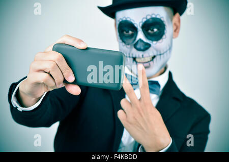 Un jeune homme avec le maquillage, le port de calaveras et noeud papillon top hat, prend un des selfies lui-même avec un smartphone Banque D'Images