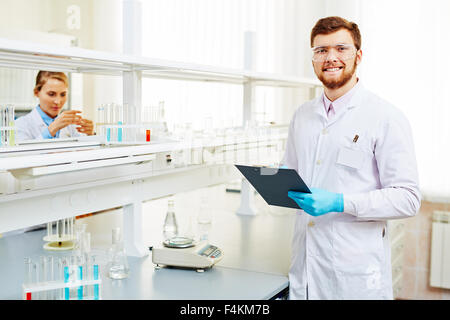 Chimiste heureux en manteau blanc, lunettes et gants de prendre des notes en laboratoire sur fond de son assistant Banque D'Images