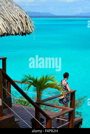 Woman sur le balcon d'un bungalow sur pilotis au toit de chaume dans un complexe de luxe avec vue sur le lagon de Bora Bora. Banque D'Images