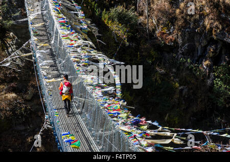 Jeune femme touristiques portant son petit enfant dans un rack sur le dos, traverser un pont suspendu avec de nombreux drapeaux de prière Banque D'Images