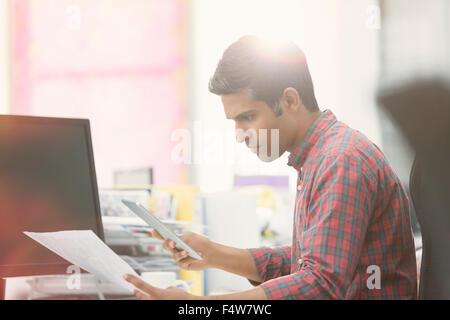 L'accent businessman with digital tablet reviewing paperwork at desk in office Banque D'Images