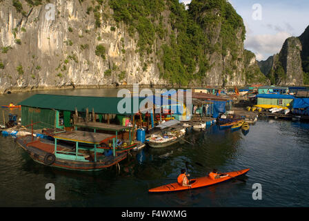 Kayak passé un village de pêcheurs flottant dans la baie d'Halong. Village de ferme du poisson entre les montagnes de calcaire karstique dans le parc national de Cat Ba, La Baie d'Halong,Ha Long, Ha Long, Halong Bay, Vietnam. Banque D'Images