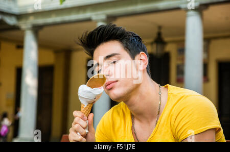 Funny jolie jeune homme assis, vêtu de T-shirt jaune tout en mangeant une délicieuse glace à la vanille à l'extérieur dans le parc, dans une guerre Banque D'Images