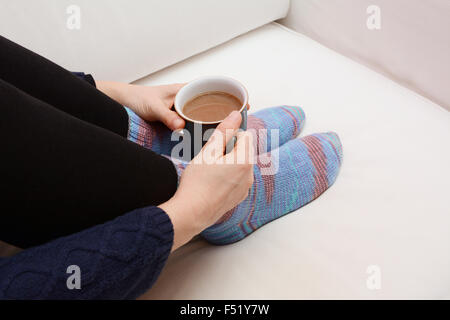 Woman's hands holding une tasse de café chaud ou un thé, recroquevillée sur le canapé, portant des chaussettes tricotées à la main confortable Banque D'Images