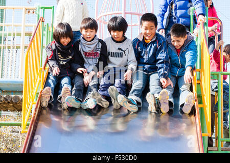 Le Japon. Cour d'école, de l'hiver. Groupe de cinq enfants, garçons, 7-8 ans, glissant sur la diapositive dans la rangée, excité, criant joyeusement. En face. La lumière du soleil. Banque D'Images