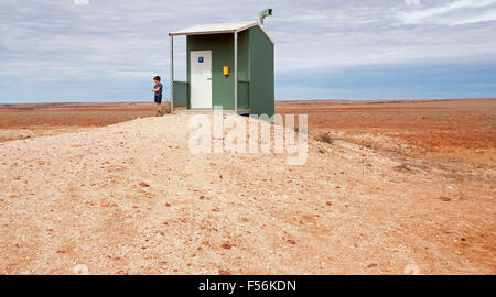 Vue humoristique de jeune garçon à côté de toilettes publiques isolées sur de vastes plaines arides de l'outback rouge en région isolée de l'Australie Banque D'Images