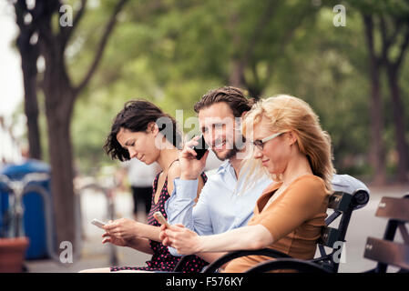 Un homme et deux femmes assises sur un banc dans un parc, la vérification de leurs téléphones portables, l'un d'effectuer un appel. Banque D'Images