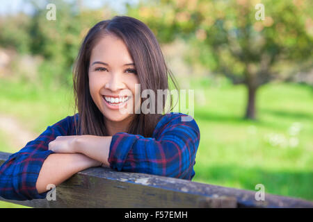 Belle jeune femme asiatique ou eurasienne girl wearing denim shirt, souriant et s'appuyant sur une clôture en sunshine Banque D'Images