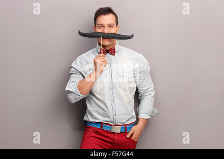 Cheerful young man holding un énorme fake moustache sous son nez et regardant la caméra Banque D'Images