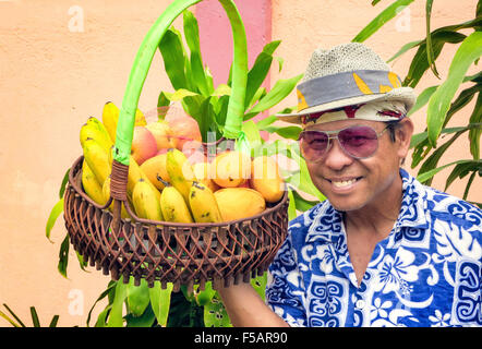 Portrait of a smiling Filipino homme portant un fedora chapeau, lunettes de soleil et une fleur shirt tenant un panier de fruits frais. Banque D'Images