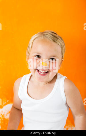 Petite fille peinture mur blanc avec la couleur orange. Elle a de la peinture orange sur son petit nez. Banque D'Images