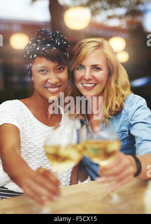 Fun young female friends avec le vin blanc de trinquer leurs verres dans un toast comme ils sourient à thecamera, mu Banque D'Images