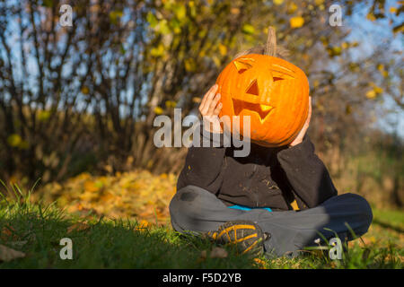 Portrait d'enfant garçon avec halloween citrouille jack-o-lanternes en face de face, assis sur l'herbe, jardin d'automne, journée ensoleillée Banque D'Images