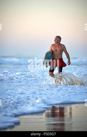 Middle aged man walking in ocean wave laver tenant une planche de surf. Tôt le matin la lumière douce sur la scène de lavage délicieux. Banque D'Images