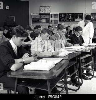 Historique, années 1950, un groupe d'étudiants en génie technique de sexe masculin assis avec leurs dossiers à un bureau dans une salle de classe avec un instructeur de sexe masculin sous un manteau blanc qui vérifie le travail d'un étudiant, Angleterre, Royaume-Uni. Banque D'Images