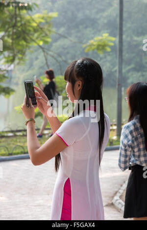 Fille vietnamienne dame en costume traditionnel ao dai, regarder l'écran sur un téléphone intelligent, se tenait près du lac Hoan Kiem, Hanoi, Vietnam Banque D'Images