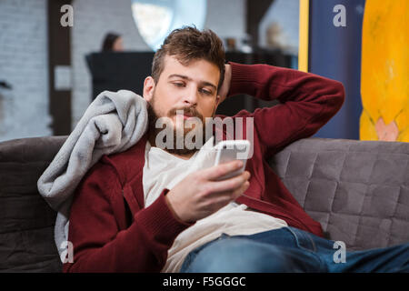 Beau jeune homme séduisant détendue couché sur le canapé gris et using cellphone Banque D'Images