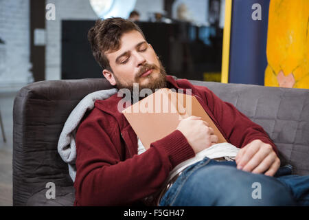 Beau barbu homme fatigué dormir sur un canapé gris holding book Banque D'Images