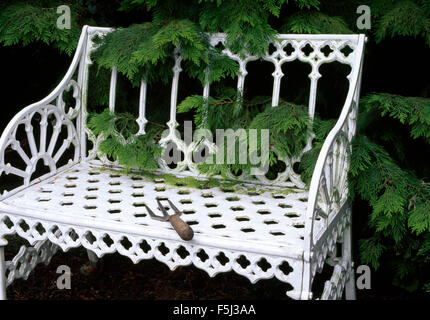 Petit jardin fourche sur un banc de jardin en fer forgé blanc en face d'un arbuste conifère vert Banque D'Images