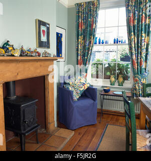 Poêle à bois dans la cheminée dans la salle à manger de style économique avec un couvercle desserré bleu sur chaise à côté de fenêtre avec des rideaux aux motifs floraux Banque D'Images