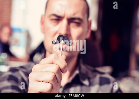 Un young caucasian man holding a fake moustache dans un bâton en face de son visage Banque D'Images